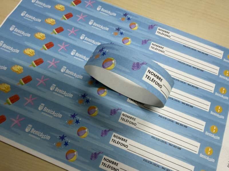 Sheets of light blue labels with fields for NOMBRE and TELEFONO and small police and beach-themed icons, a roll of labels resting on top.
