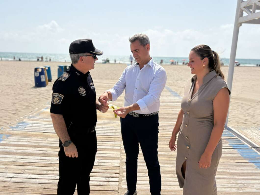Police officer on a beach boardwalk accepts a small item from a man in a white shirt while a woman watches on.