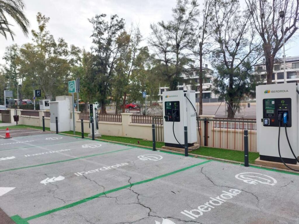 Row of Iberdrola electric vehicle charging kiosks along a sidewalk in a parking lot with green EV bays.