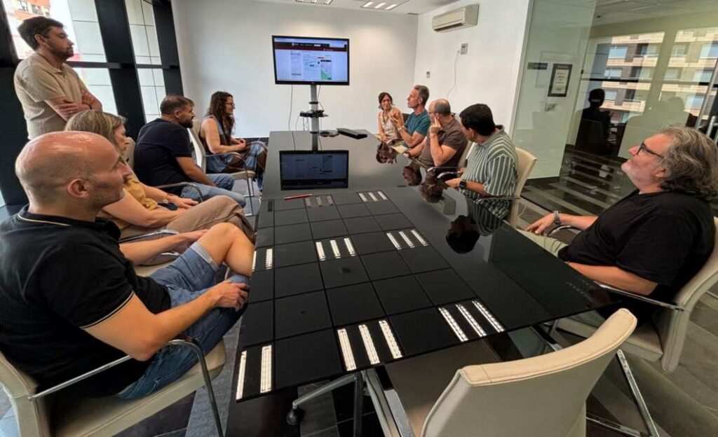 Team of ten people in a modern conference room watching a presentation on a wall-mounted monitor on display.