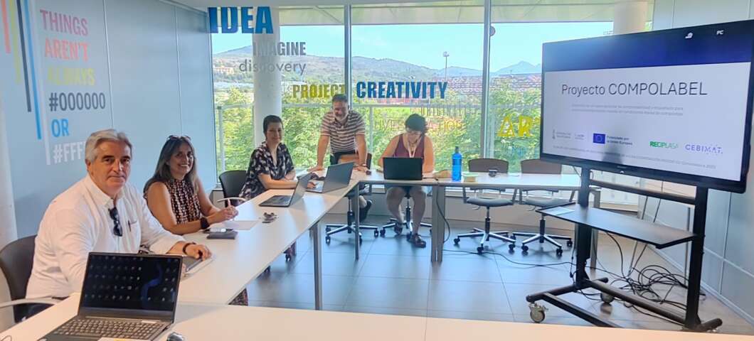 Six colleagues sit around a white meeting table in a bright glass-walled office; a large monitor shows 'Proyecto COMPOLABEL' during a presentation.