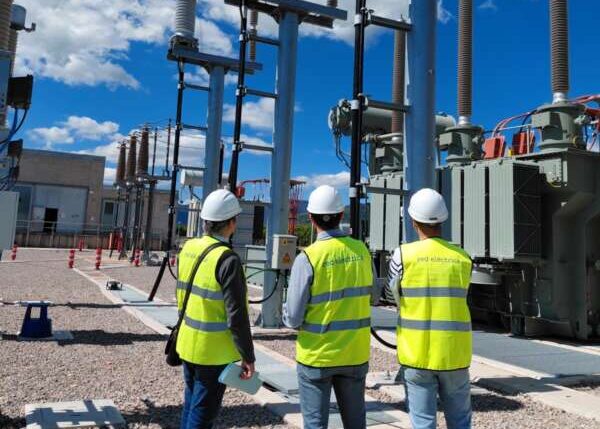 Three workers in yellow safety vests and white hard hats inspect tall high-voltage equipment at a substation under a blue sky.