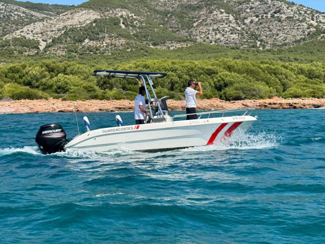 White coast-guard boat with two crew, one using binoculars, on blue sea near a rocky, tree-covered coast.