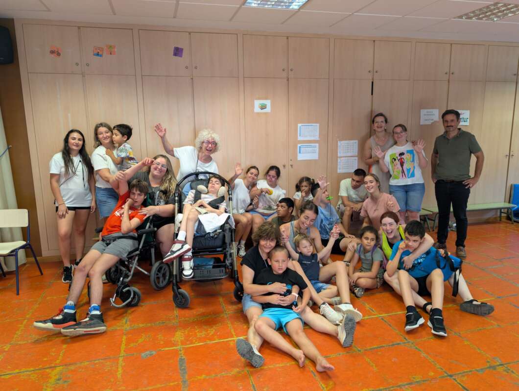 Group photo of children and adults in a classroom-like room, many sitting on the floor and two in wheelchairs, all smiling and waving.