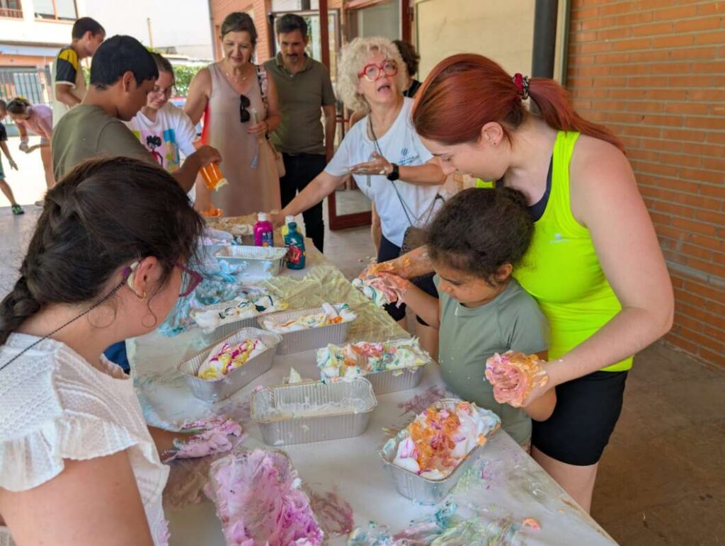 People at a long table covered in paint, helping children make colorful crafts with gooey paint on their hands outside a brick building