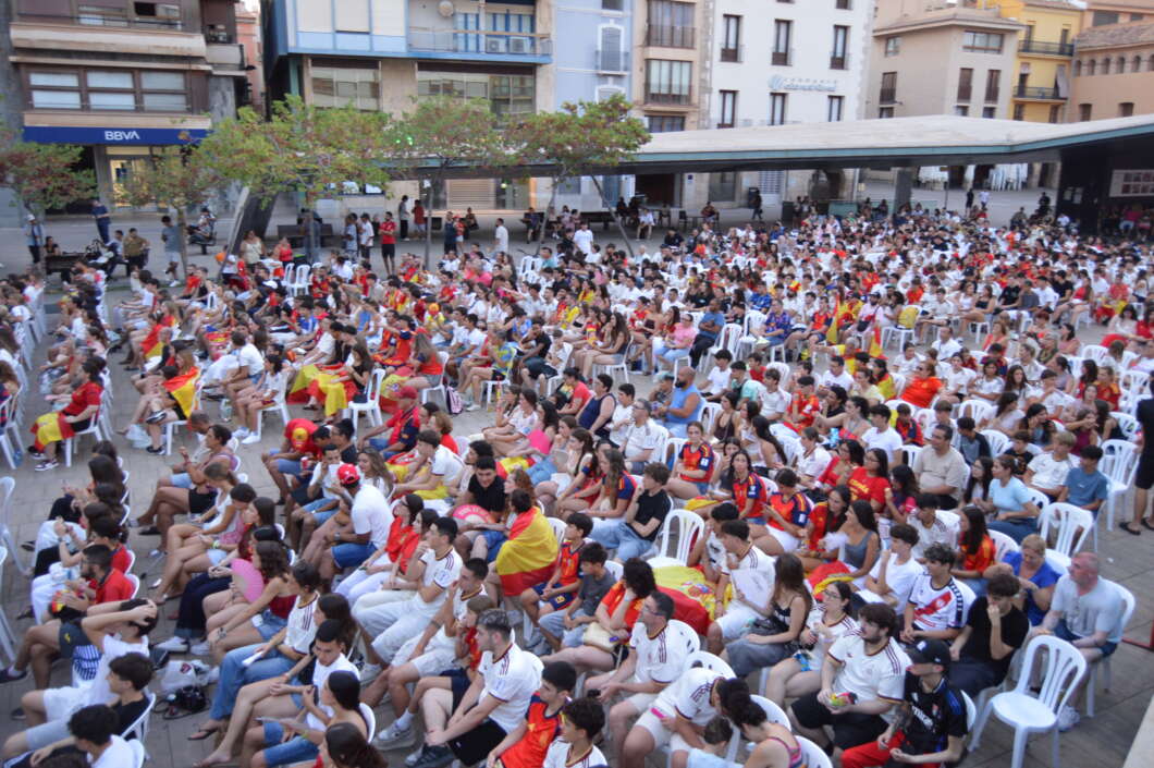 Large crowd of people seated on white chairs in an outdoor plaza, many wearing red and yellow Spain flags and colors.