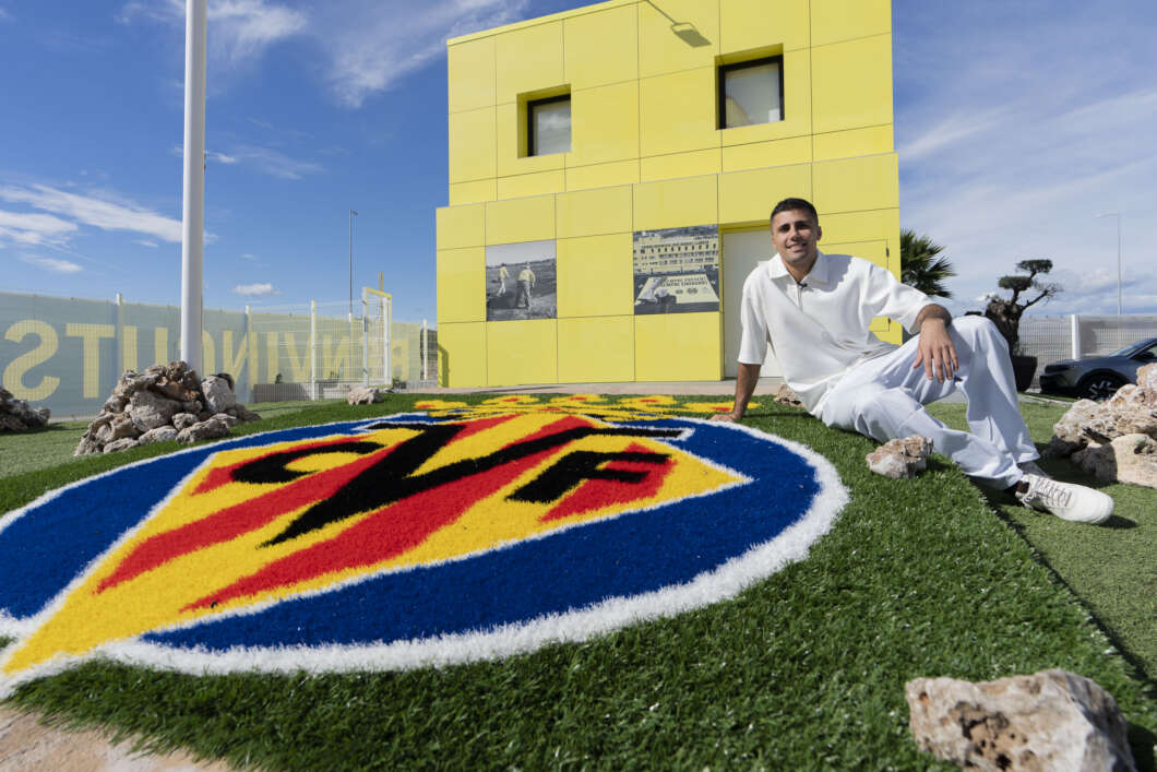 Man in white outfit sits on green turf beside a large circular blue‑red‑yellow logo in front of a bright yellow building on a sunny day.