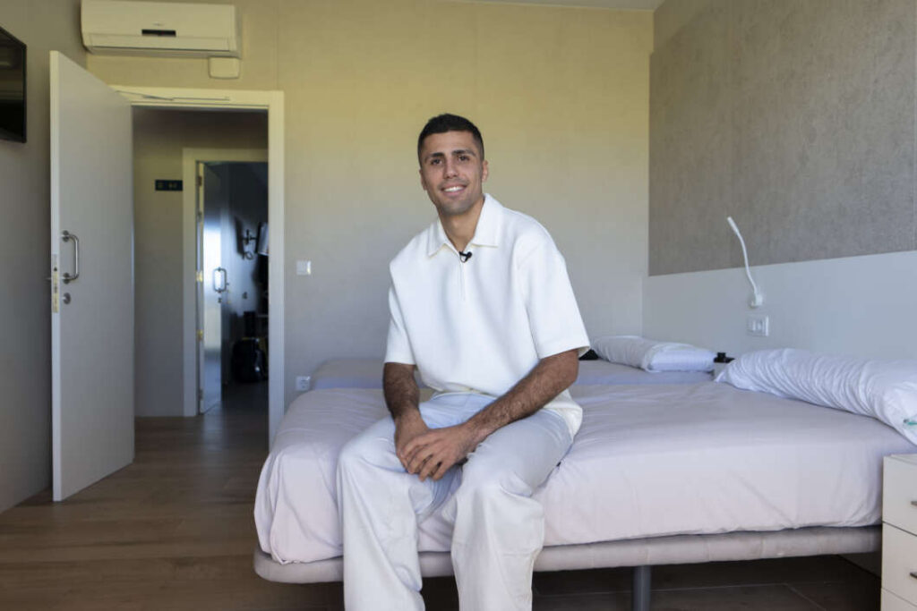 Smiling man in a white polo sits on the edge of a made hotel bed in a modern room with an open door beside him.