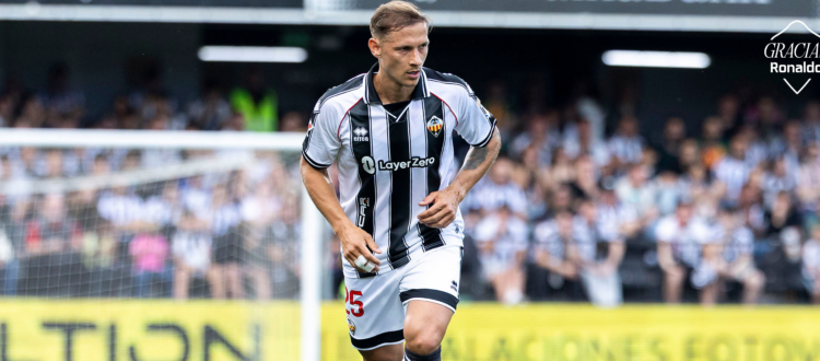 Soccer player in Valencia CF black-and-white striped kit runs toward the ball during a match, with fans in the stands behind.
