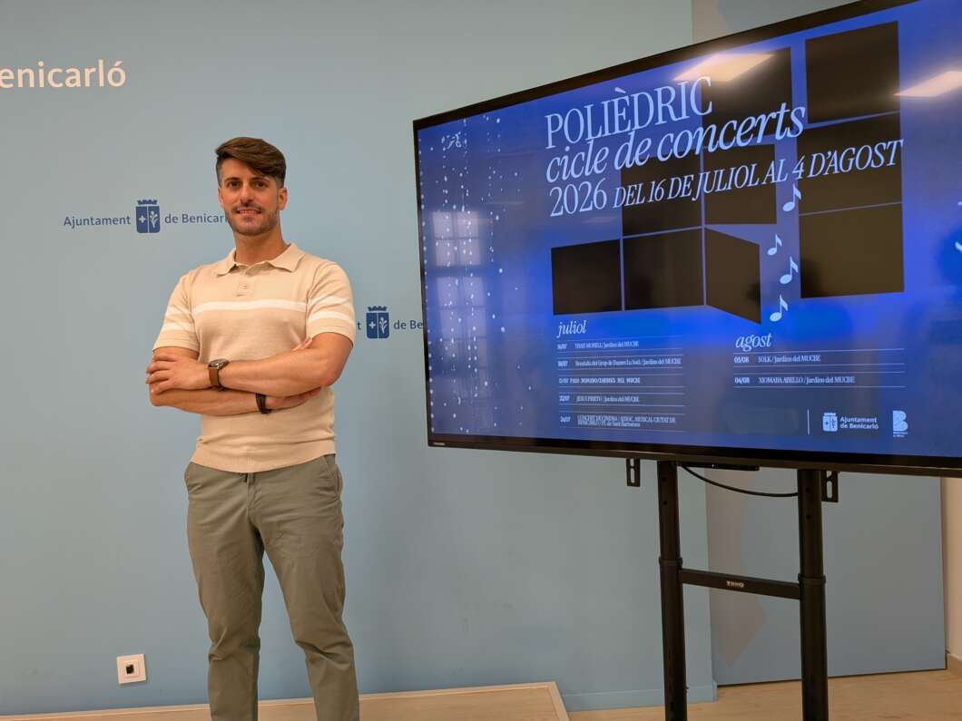Man stands with arms crossed beside a tall screen displaying a blue poster for a 2026 concert cycle event while a light-blue wall sits behind him.