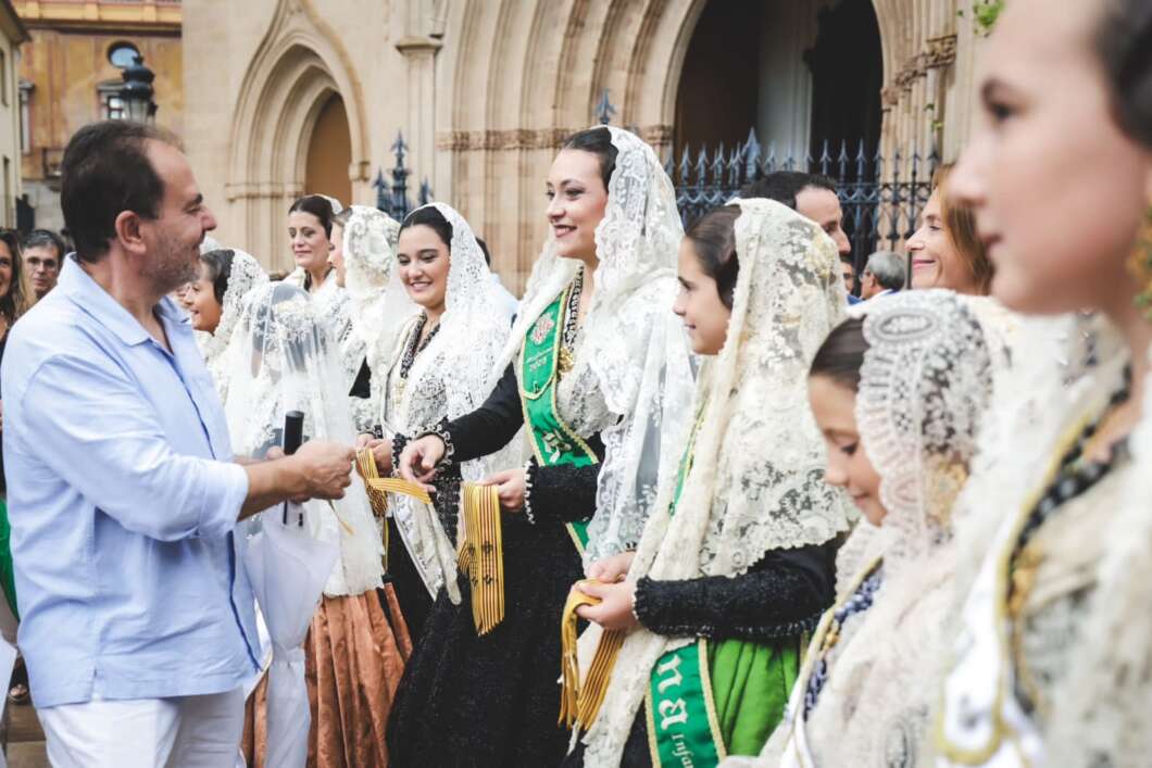 Man in a light blue shirt hands ceremonial sashes to a line of women in white lace mantillas outside a historic building, smiling.