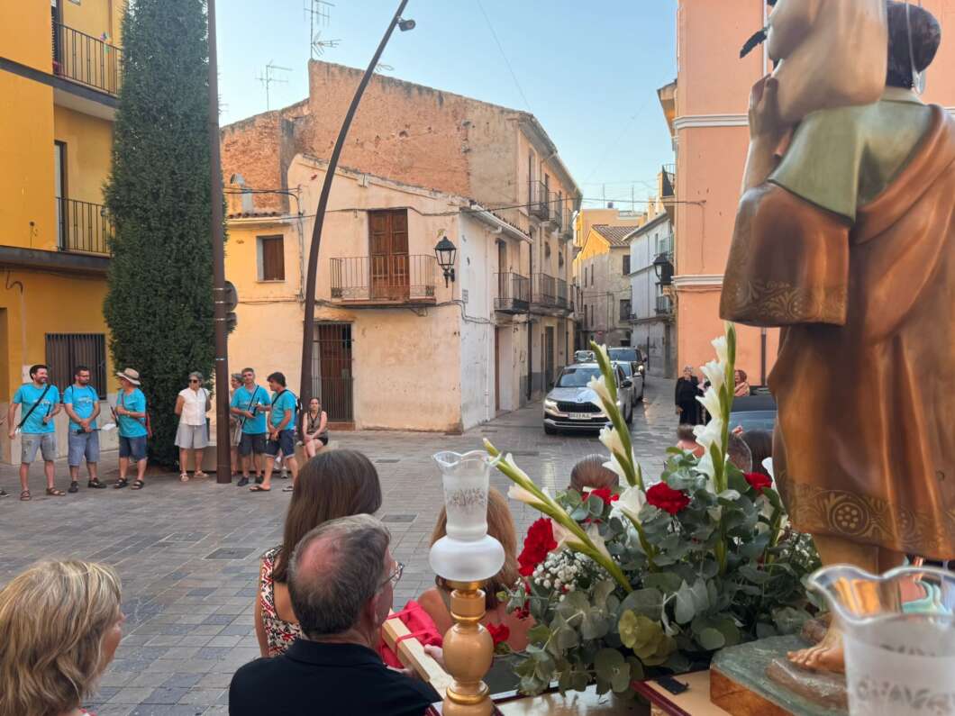 Statue on a flower-decorated float́ passes through a cobblestone street as onlookers and volunteers in blue shirts watch from the sidewalk.