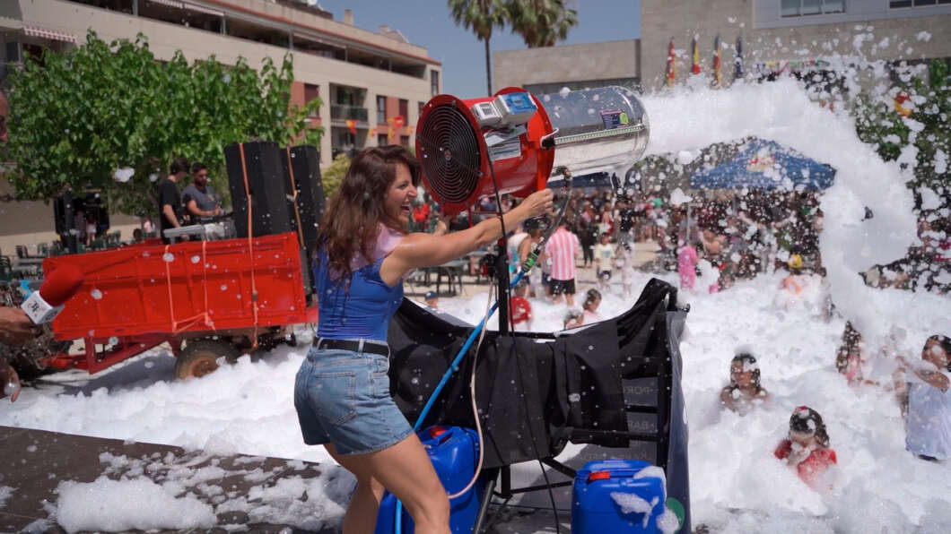 Woman in a blue top and denim shorts operates a foam cannon, spraying foam over a crowded outdoor foam party.
