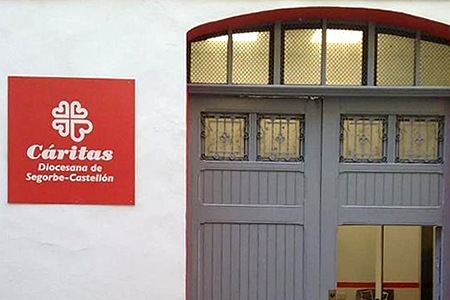 Red Cáritas sign with logo and 'Diócesis de Segorbe-Castellón' on a white wall beside a gray double-door entry.