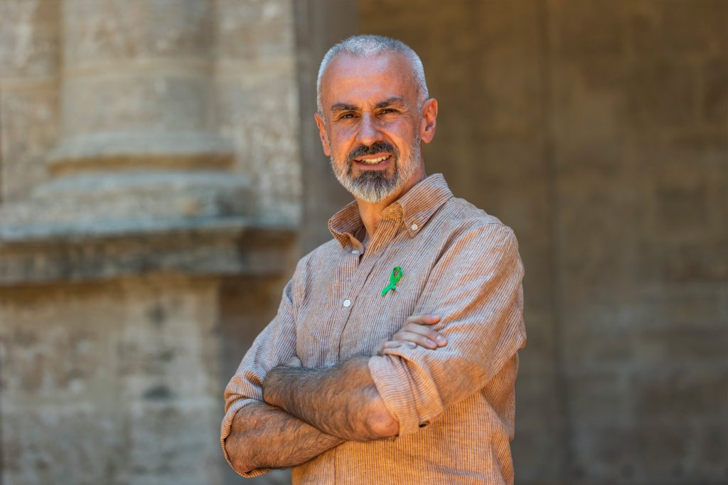 Smiling man with gray beard in a light plaid shirt, arms crossed, wearing a green awareness ribbon against a stone backdrop.