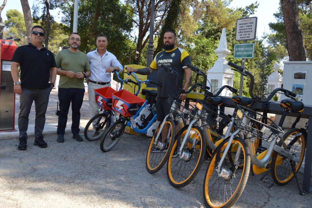 Group of four men standing next to a row of yellow-wheeled rental bikes in a park. Two bikes have red baskets in front; the men appear to be posing with the bikes under a sunny, tree-lined area.