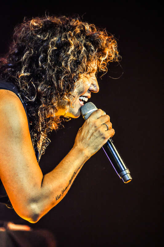 Singer with curly hair performing on stage, gripping a handheld microphone and singing passionately.