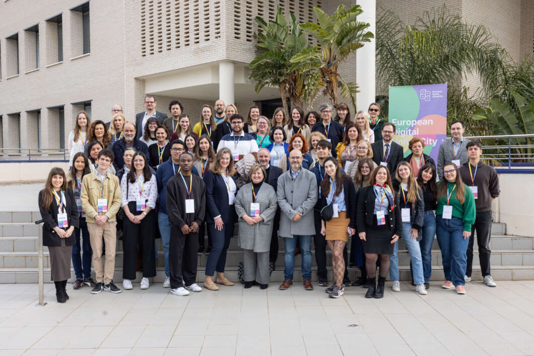 Group of diverse people wearing badge lanyards pose on steps outside a beige building, with a European Digital banner in the background.
