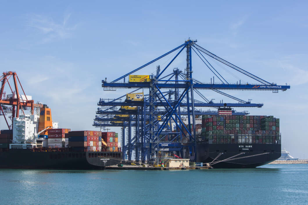 Container ship at a busy port with stacked colorful containers and blue gantry cranes overhead.