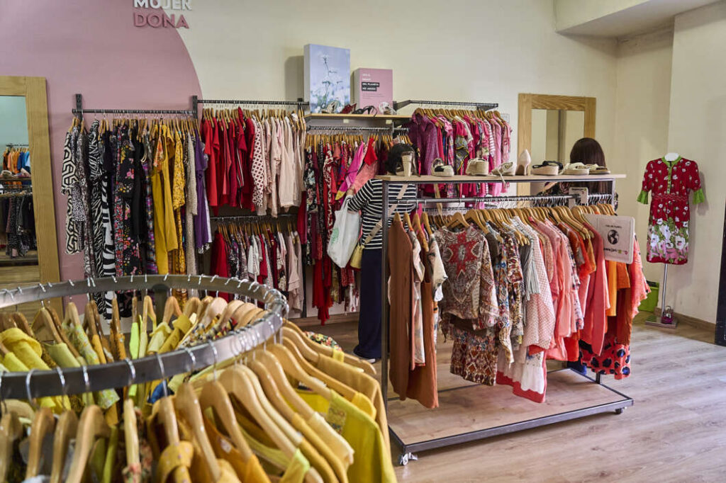 Bright women’s clothing store with racks of colorful dresses, tops, and accessories; a mannequin in a red dress on the right and shelves with hats above the racks.