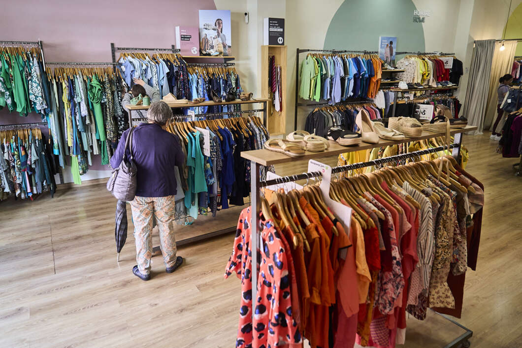 Shopper browsing racks of colorful clothing in a bright clothing store, with hats on a display table nearby.