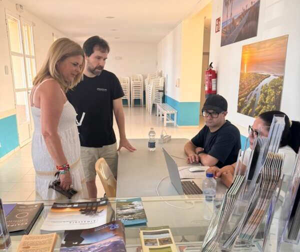 Woman in a white dress and a man in a black shirt stand at a reception desk, talking to two staff members with laptops behind the glass counter; travel brochures are spread across the desk in a bright info center.