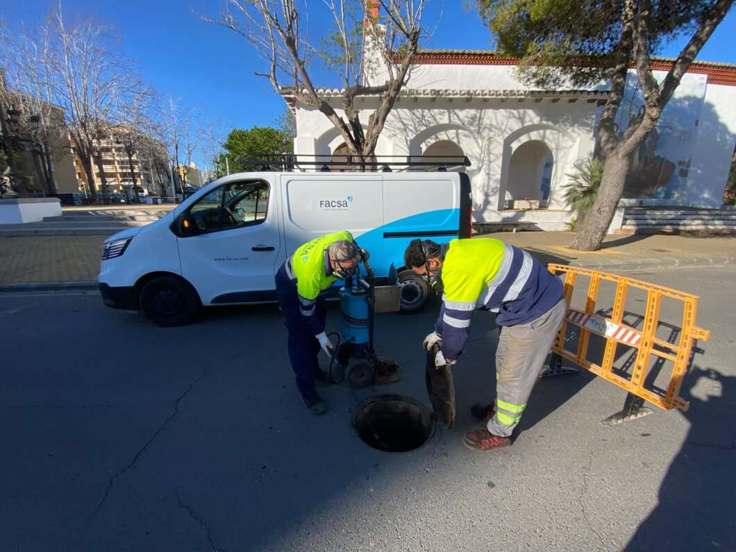 Two workers in high‑visibility jackets inspect and work inside an open manhole on a city street, as a white utility van labeled 'Facsa' sits nearby.