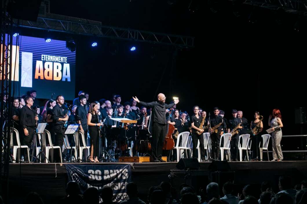 Conductor leads a large musical ensemble on stage with singers and instrumentalists, a banner for ABBA in the background.