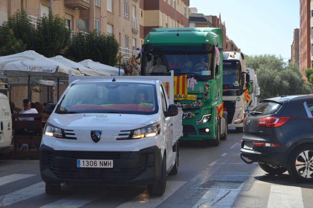 White parade van leads a procession of decorated trucks along a sunlit urban street, with pedestrians seated at a sidewalk cafe nearby.