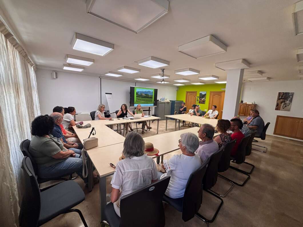 A group of adults sits around a U-shaped arrangement of tables in a meeting room, watching a presentation on a wall-mounted screen.