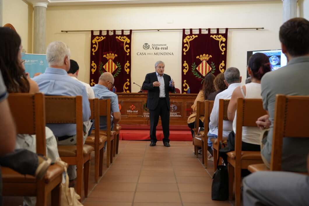 Speaker in a suit presenting at a podium in a conference room, audience seated, with burgundy banners and crest behind him on stage behind glass doors.