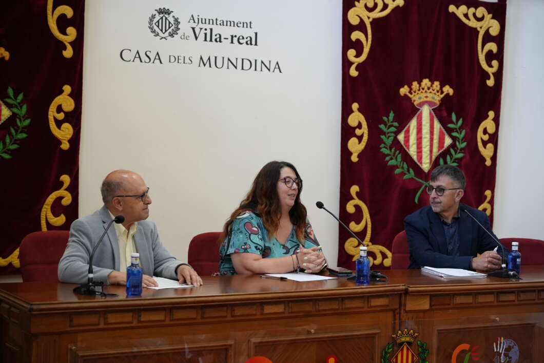 Panel discussion at Vila-real city hall with three speakers at a wooden table and ceremonial banners behind them.