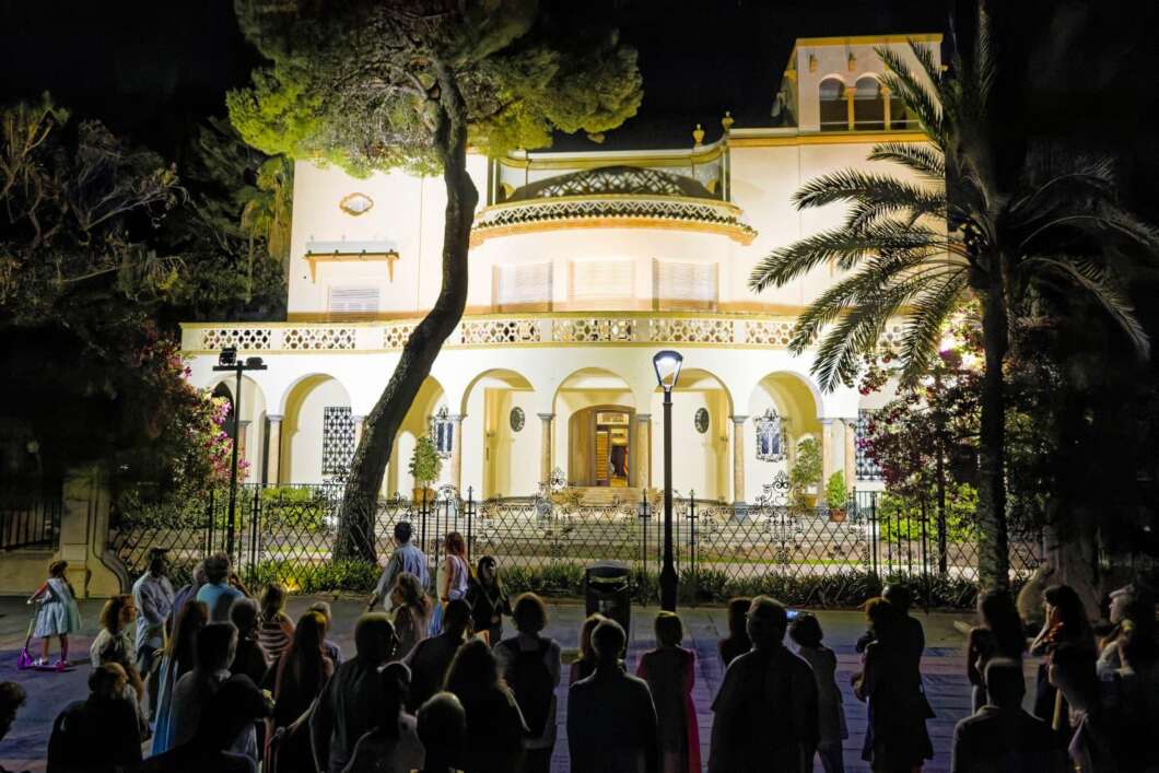 Night scene of a well-lit Mediterranean villa with arches, palm trees, and a crowd gathered outside behind a fence.