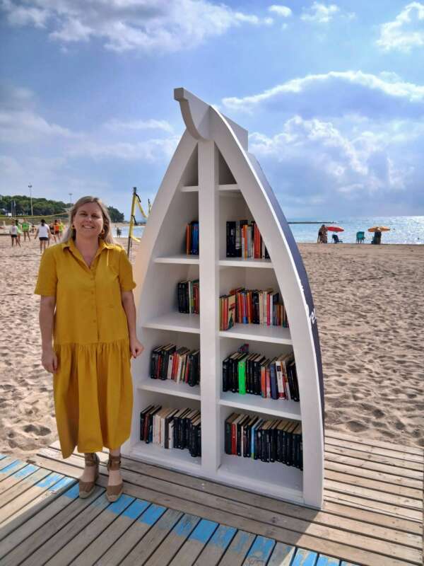 A woman in a yellow dress stands beside a white, boat-shaped bookshelf filled with books on a sandy beach.
