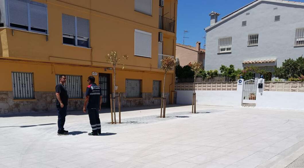 Two men stand and talk in a sunny courtyard beside a yellow building, one in casual dark clothes and the other in a reflective-work uniform.