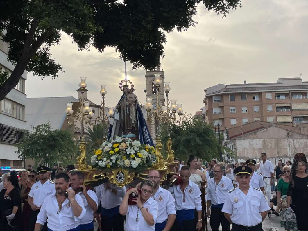 Religious procession on a city street: a gilded floral float carries a statue of the Virgin Mary, surrounded by white and yellow flowers, carried by men in white shirts while crowds walk behind at dusk.