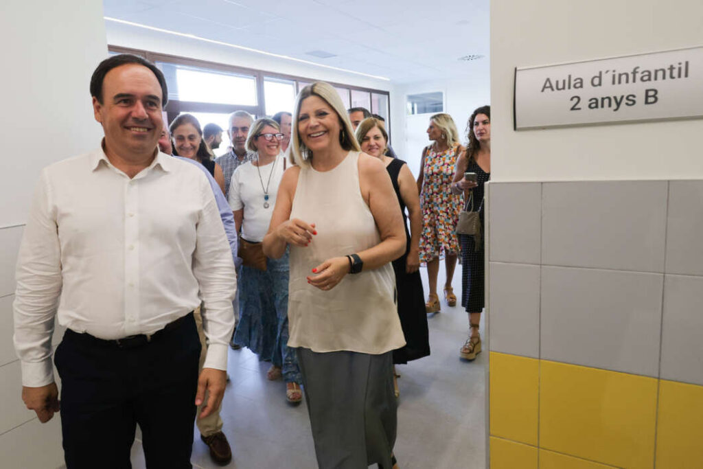 Smiling group of adults walking down a school hallway toward a classroom door with a Catalan sign reading 'Aula d'infantil 2 anys B' on the wall.