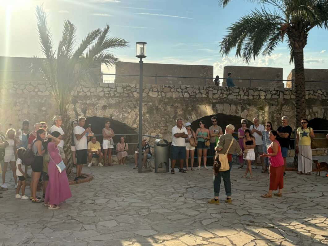 Tour group gathered in a sunny stone plaza, listening to a guide near a historic stone wall and palm trees.