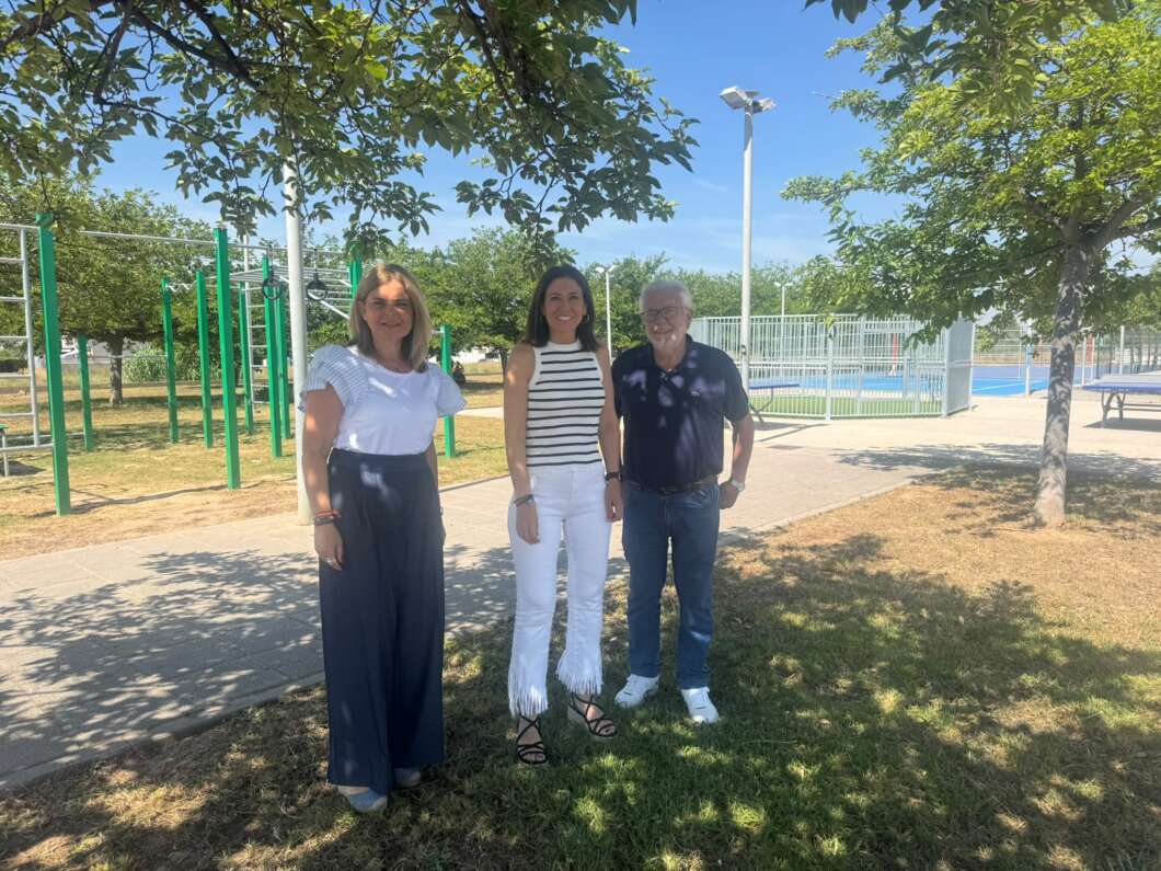 Three adults posing together in a sunny park near outdoor exercise equipment and a fenced pool in the background.